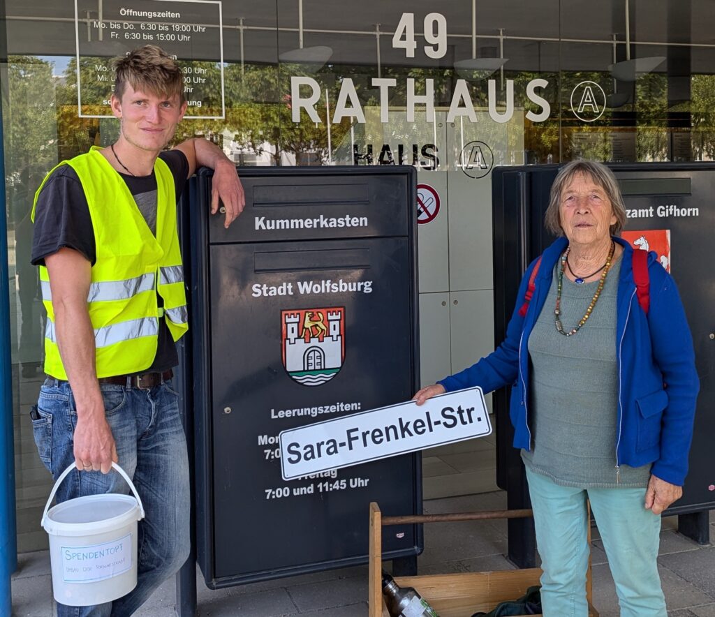 Zwei Menschen mit einem Straßenschild Sara-Frenkel-Straße vor einem großen Kummerkasten vor einem Rathaus.